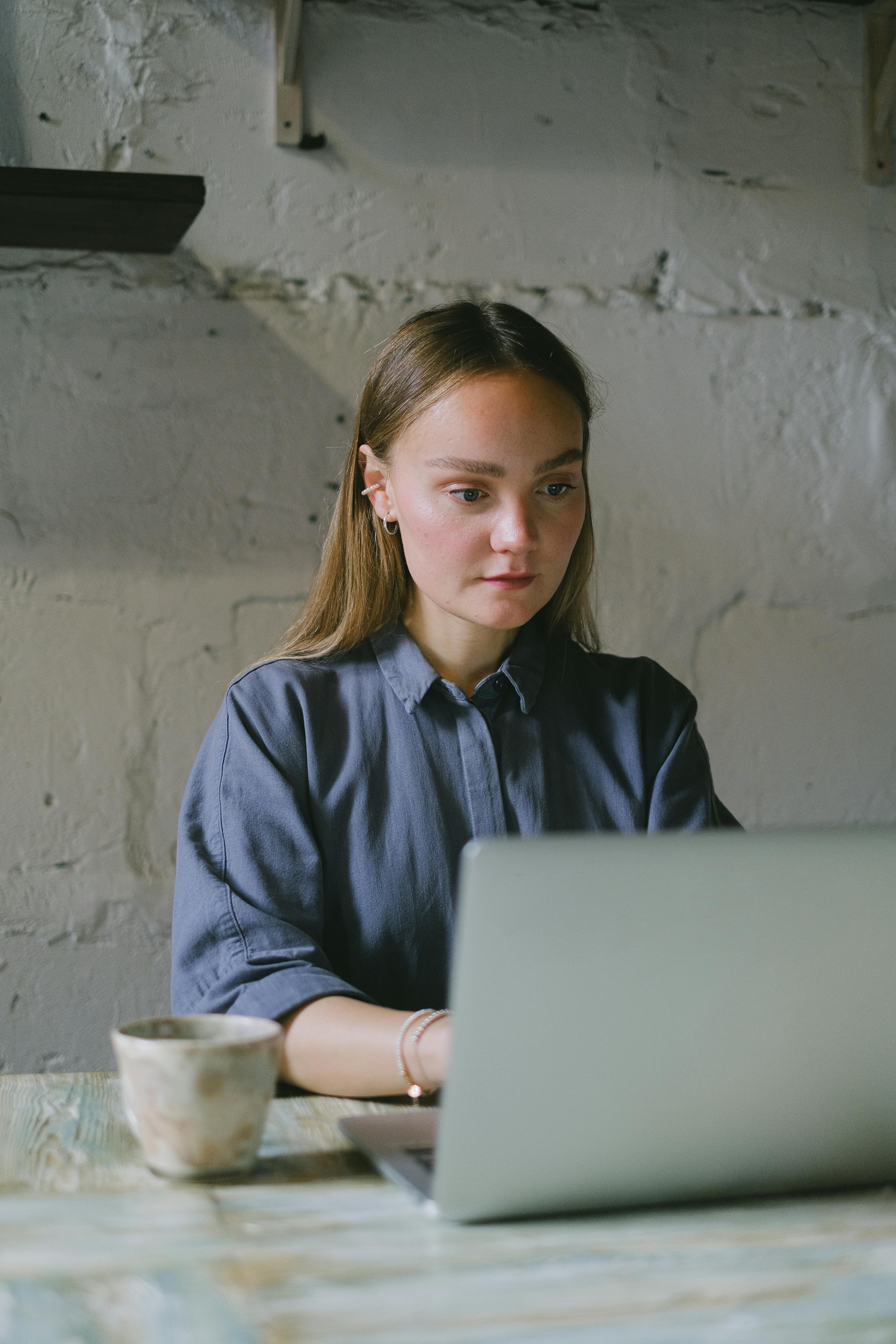 Serious young woman in casual clothes sitting at table and working remotely on netbook near mug with drink in workplace